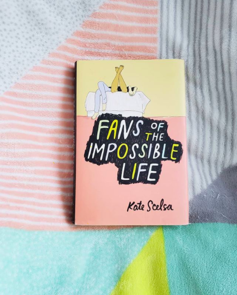 Paperback of "Fans of the Impossible Life" by Kate Scelsa atop a multi-patterned fuzzy blanket
