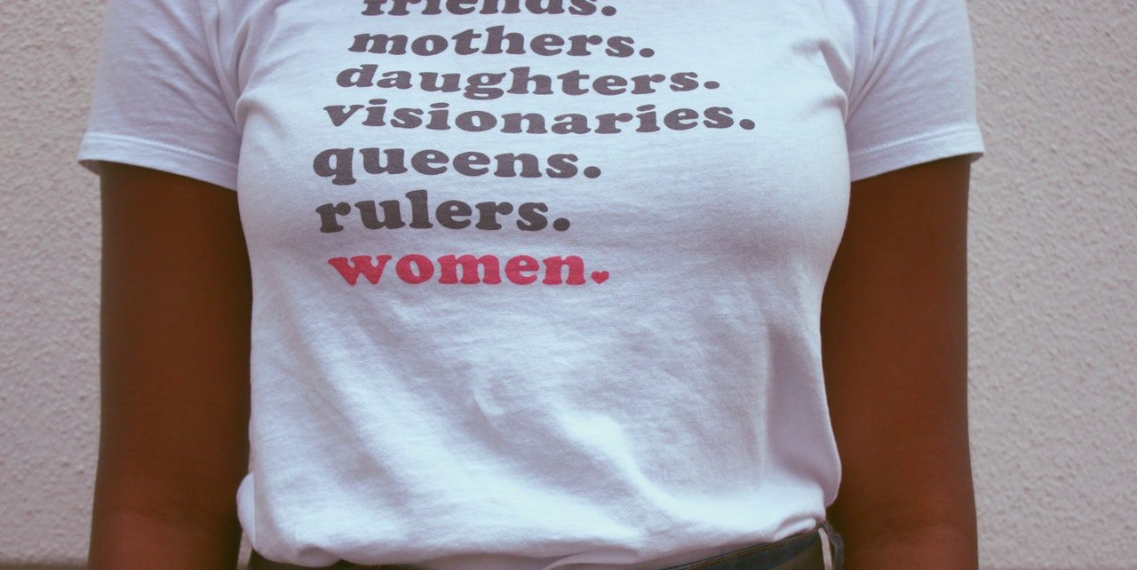 A woman wearing a white t-shirt reading "friends. mothers. daughters. visionaries. queens. rulers." in black, followed by "women" and a tiny heart in place of a period in bright pink